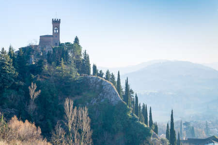 A  medieval crenellated clock tower stands on a peak overlooking the valley of a country of farmland, bushes, cypresses and other tress in the mist of a sunny winter dayのeditorial素材