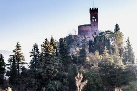 A  medieval crenellated clock tower stands on a peak overlooking the valley of a country of farmland, bushes, cypresses and other tress in the mist of a sunny winter dayのeditorial素材