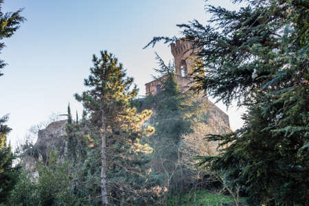 A  medieval crenellated clock tower stands on a peak overlooking the valley of a country of farmland, bushes, cypresses and other tress in the mist of a sunny winter dayのeditorial素材