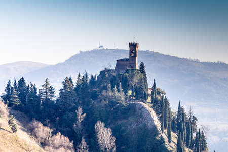 A  medieval crenellated clock tower stands on a peak overlooking the valley of a country of farmland, bushes, cypresses and other tress in the mist of a sunny winter dayのeditorial素材