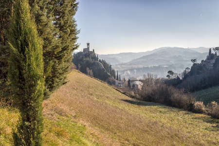 A  medieval crenellated clock tower stands on a peak overlooking the valley of a village and a country of farmland, bushes, cypresses and other tress in the mist of a sunny winter dayのeditorial素材