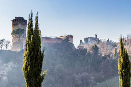 Cypress trees bordering a walk path to a medieval fortress and a sanctuary with steeple on misty hills in a contryside of bushes and cypress trees in a winter sunny dayのeditorial素材