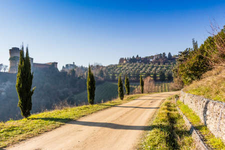 Cypress trees bordering a walk path to a medieval fortress and a sanctuary with steeple on misty hills in a contryside of bushes and cypress trees in a winter sunny dayのeditorial素材