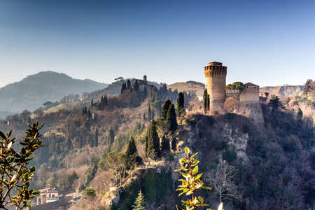 The brick walls of a medieval fortress and a sanctuary with steeple devoted to Blessed Virgin Mary on misty hills in a countryside of bushes and cypress trees in a winter sunny dayのeditorial素材