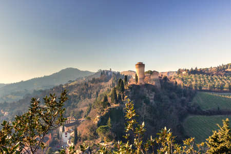 The brick walls of a medieval fortress and a sanctuary with belfry on misty hills in a countryside of cultivated fields, bushes and cypress trees in a winter sunny dayのeditorial素材
