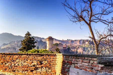 The brick walls of a medieval fortress and a sanctuary with steeple devoted to Blessed Virgin Mary on misty hills in a countryside of bushes and cypress trees from bulwark in a winter sunny dayのeditorial素材
