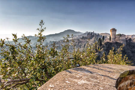 The brick walls of a medieval fortress and a sanctuary with steeple devoted to Blessed Virgin Mary on misty hills in a countryside of bushes and cypress trees from bulwark in a winter sunny dayのeditorial素材