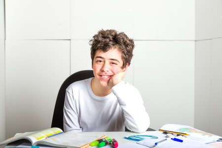 Happy Caucasian smooth-skinned boy holds his chin with his left hand resting his elbow on the desk...
