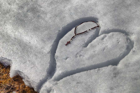 Wooden Rosary beads left on a heart drawn by hand in the fresh snowの写真素材