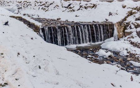 mountain stream in the middle of snow and rocks along banks of roots and weeds on Dolomites mountainsの写真素材