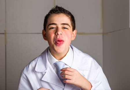 Boy dressed as a doctor in light blue tie and white coat helps to feel medicine more friendly: he is showing his tongue with a pill on it. His acne skin has not ben retouchedの写真素材