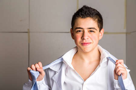 Pimply but handsome boy doctor tired but serene while undressing: he is untying the knot of the light blue tie and opening his white shirt. His acne skin has not ben retouchedの写真素材