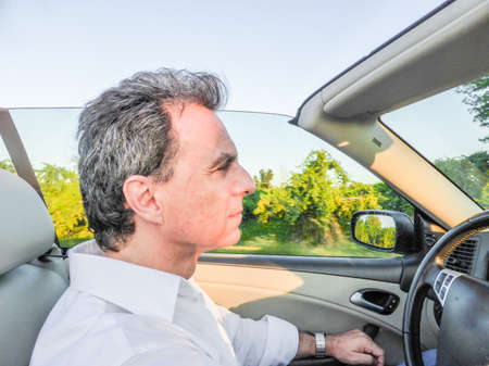 Side view of handsome middle-aged man with salt pepper hair and wearing a white shirt while he is driving a convertible car in the countrysideの写真素材