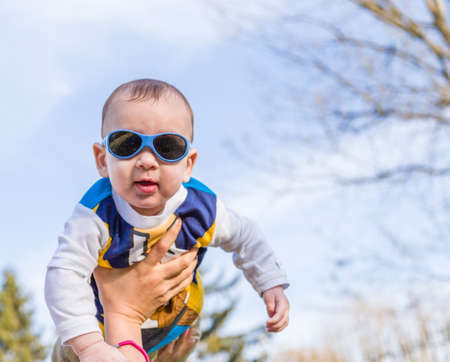 Cute 6 months old baby with Light brown hair in white, blue and brownish long-sleeved shirt wearing blue googles is raised in the air, embraced and held by his mumの写真素材
