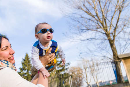 Cute 6 months old baby with Light brown hair in white, blue and brownish long-sleeved shirt wearing blue googles is raised in the air, embraced and held by his mumの写真素材