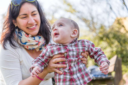 Cute 6 months old baby with Light brown hair in red checkered shirt and beige pants is embraced and held by his smiling Hispanic mummyの写真素材