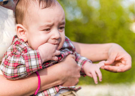 Cute 6 months old baby with Light brown hair in red checkered shirt and beige pants is biting his fingers while embraced by his Hispanic motherの写真素材