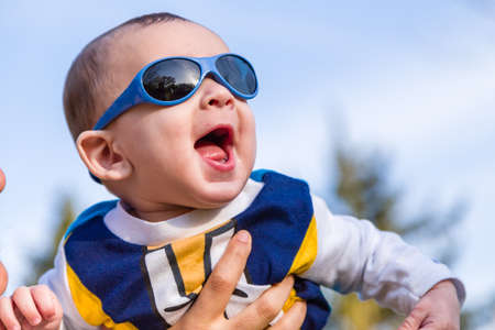 Cute 6 months old baby with Light brown hair in white, blue and brownish long-sleeved shirt wearing blue goggles is embraced and held by his mum: he seems very happy and smilesの写真素材