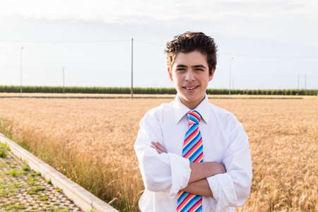 Handsome Caucasian boy wearing a white shirt and a regimental tie with red, fuchsia, orange, blue, indigo and white stripes is crossing arms on a field of golden wheat ears at sunsetの写真素材