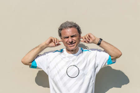 Handsome middle-aged man with salt pepper hair dressed with sports shirt is plugging his ears with fingers to not hear noise or words: he seems very disturbedの写真素材