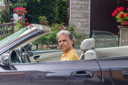 Side view of classy senior sportsman with three-day beard and salt cand pepper hair wearing a yellow polo shirt while he is driving a dark brown car in residential neighborhoodの写真素材