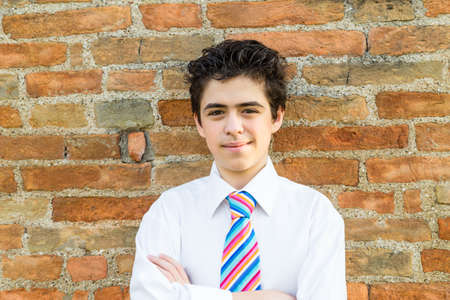 Handsome Caucasian boy wearing a white shirt and a regimental tie with red, fuchsia, orange, blue, indigo and white stripes is crossing his arms in front of a brick wall at sunsetの写真素材