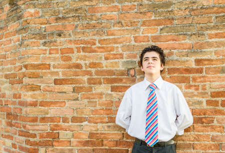 Handsome Caucasian boy leaning against an ancient brick wall while he is wearing a white shirt and a regimental tie with red, fuchsia, orange, blue, indigo, blue and white stripesの写真素材