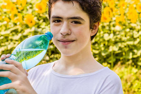 Environmentally-friendly living - Caucasian boy in a field of sunflowers is about to drink clear water from a plastic bottle green bluish to quench thirst on a warm and bright summer dayの写真素材