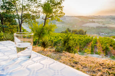dinner in the country - glass of clear, fresh and sweet water on a table with vineyards, farmlands and green vegetation of the countryside in the backgroundの写真素材