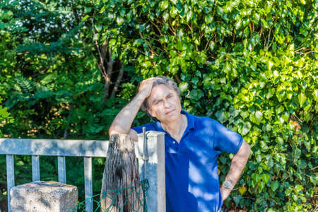 Caucasian middle-aged man, elegant and sporty dressing a blue polo, has a thoughtful look while holding head and leaning against a steel gate in the Italian countryside in summerの写真素材
