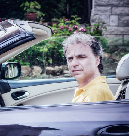 Side view of classy  40 years old sportsman with three-day beard and salt and pepper hair wearing a yellow polo shirt while he is driving a dark brown car in residential neighborhoodの写真素材