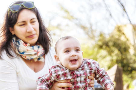 Cute 6 months old baby with Light brown hair in red checkered shirt and beige pants is embraced and held by his smiling Hispanic mummyの写真素材
