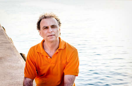 Classy  40 years old sportsman with three-day beard and salt and pepper hair wearing an orange polo shirt while he is sitting on the pier and restingの写真素材