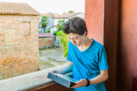 Latin boy lost in thought as his eyes look away toward the tablet PC with a red wall and green leaves as backgroundの写真素材