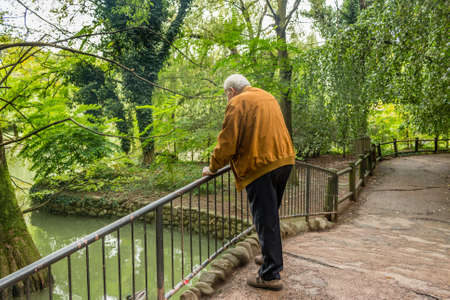 senior with suede jacket and white shirt in a green park leaning against a railing watching a pond