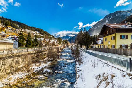 River passing through snowy alpine village in Italy illuminated by sun with mountains in the backgroundの写真素材