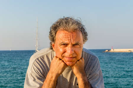 Smiling and tanned middle-aged Caucasian man relaxing on vacation, holding his chin in front of the breathtaking panorama of an Italian seaside townの写真素材
