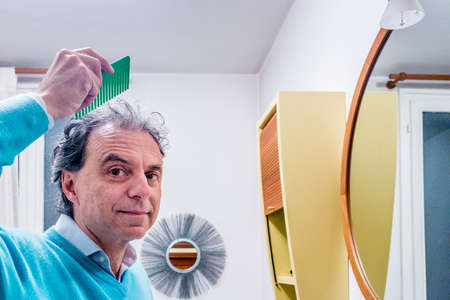balding fifty man combing his hair in the bathroom before the mirrorの写真素材