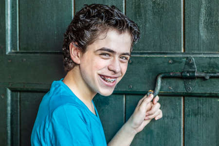 smiling teenager shows braces while moving the bolt of an old wooden doorの写真素材