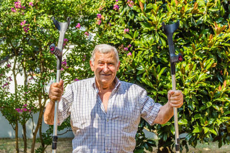 Funny faces of elderly octogenarian male triumphantly holding crutches on the patio of the houseの写真素材