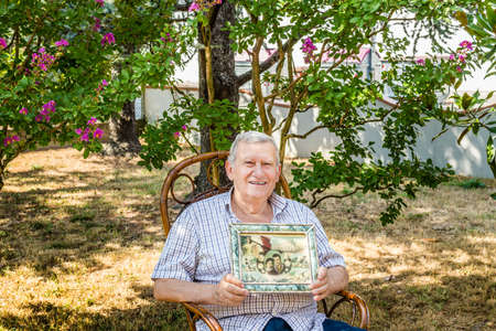 elderly octogenarian male smiling and showing old vintage photo in a frame while sitting on wicker chair in the gardenの写真素材