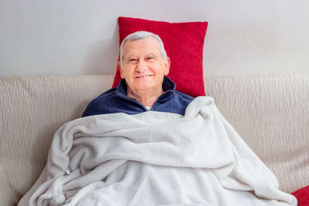 senior is smiling while resting on the couch covered with a soft white blanket and red cushionsの写真素材