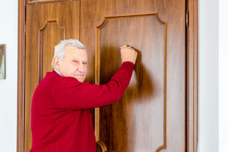 eighty-year-old man in red sweater knocks on the wooden door outsideの写真素材