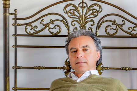 middle-aged man with piercing green eyes leaning against the bed headboard of wrought iron looks intensively and unfriendlyの写真素材