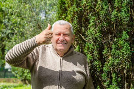 Elderly Caucasian in green garden makes the sign of satisfaction or success by raising the thumbの写真素材