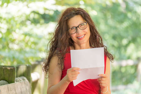 gorgeous middle-aged woman in a red dress and long brown wavy hair is reading a paper and smiling while wearing a pair of nerdy eyeglasses in a gardenの写真素材