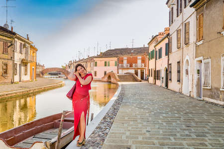 environmental portrait of sexy woman in red tight dress walking along the water canals in the hamlet of an old village in Italy, Comacchio, known as Little Veniceの写真素材