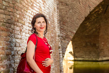 environmental portrait of sexy woman in red tight dress standing against the walls of an old bridge in the hamlet of an old Italian village, Comacchio, known as Little Veniceの写真素材