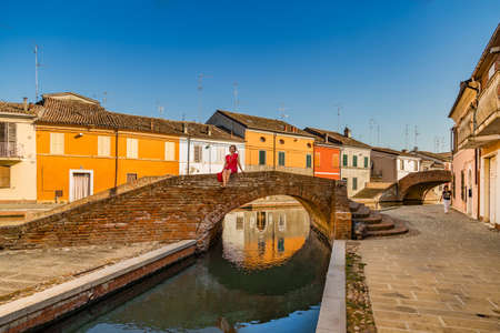middle-aged woman among colorful historical houses sitting on an ancient brick bridge on a water canal of a lagoon town in Northern Italyの写真素材