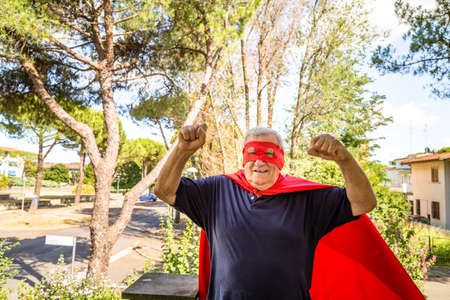 Funny and smiling senior man posing as superhero with red cape and mask is showing muscles raising arms in a quiet residential neighborhoodの写真素材